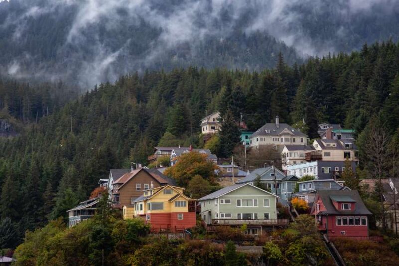 Een schilderachtig uitzicht op de stad Ketchikan in Alaska, met kleurrijke huizen die tegen een heuvel aanleunen, omringd door dichtbeboste bergen. De mist hangt laag boven de bomen, wat de scène een mysterieuze sfeer geeft.