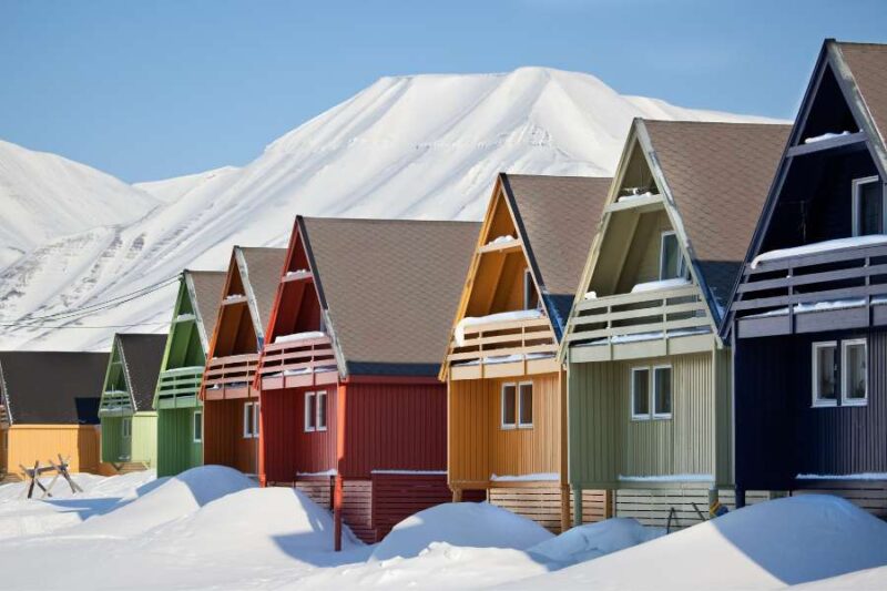 Een rij kleurrijke houten huizen in Longyearbyen, Svalbard, Noorwegen, staan in de sneeuw. De huizen hebben steile daken en zijn in verschillende kleuren geschilderd, terwijl op de achtergrond besneeuwde bergen zichtbaar zijn onder een heldere hemel.