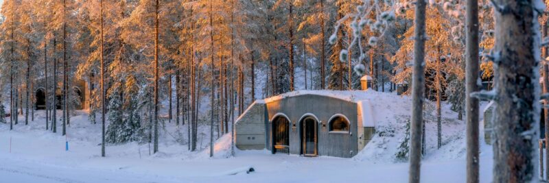 Kaamos Lodge cabin in de sneeuw met zonnestralen door de bomen.